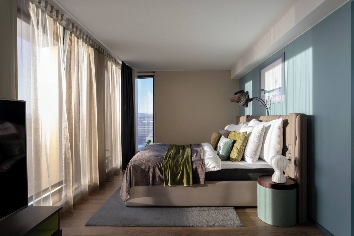 A modern bedroom with a bed, curtains, a TV, and a city view.