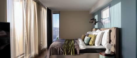 Un dormitorio moderno con cama, cortinas, televisor y vista a la ciudad.