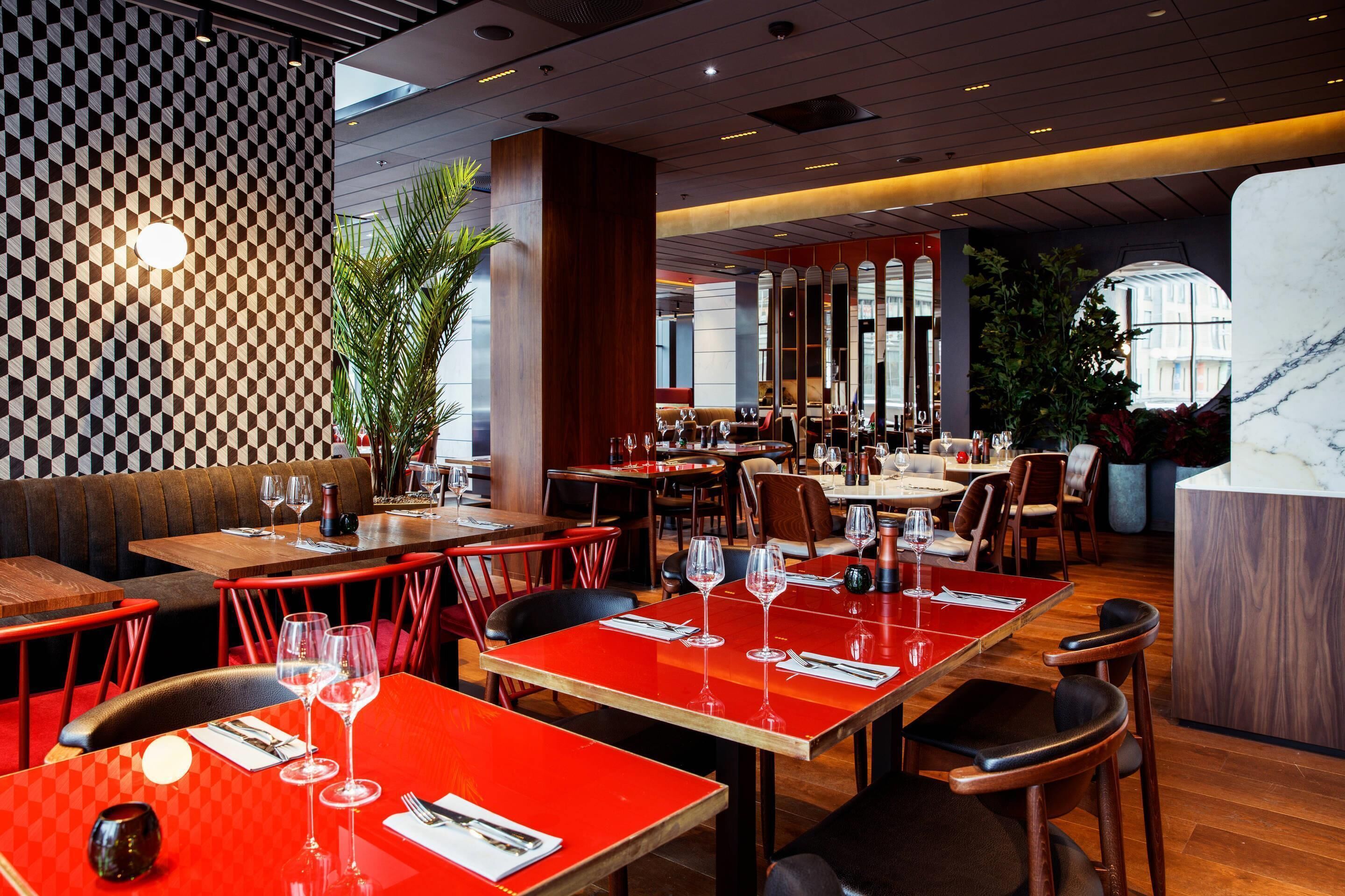 Restaurante com mesas vermelhas, cadeiras de madeira e parede estampada.