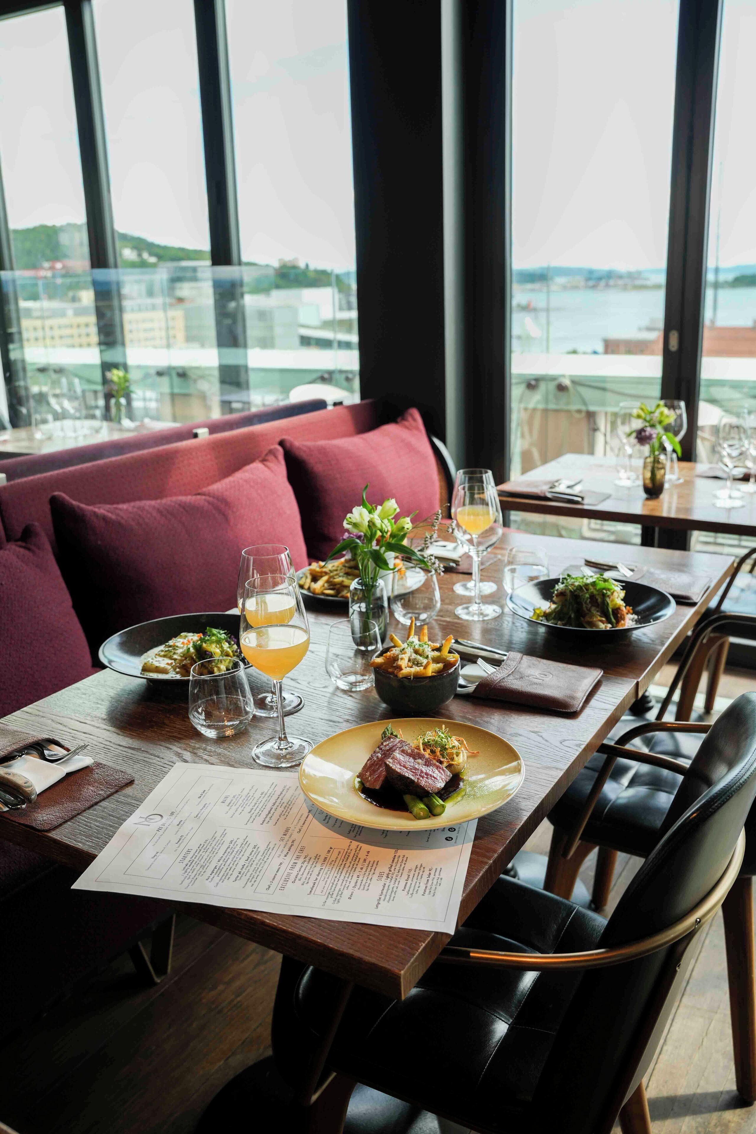 A restaurant with a wooden table set for dining, a menu, and a view of the city through large windows.