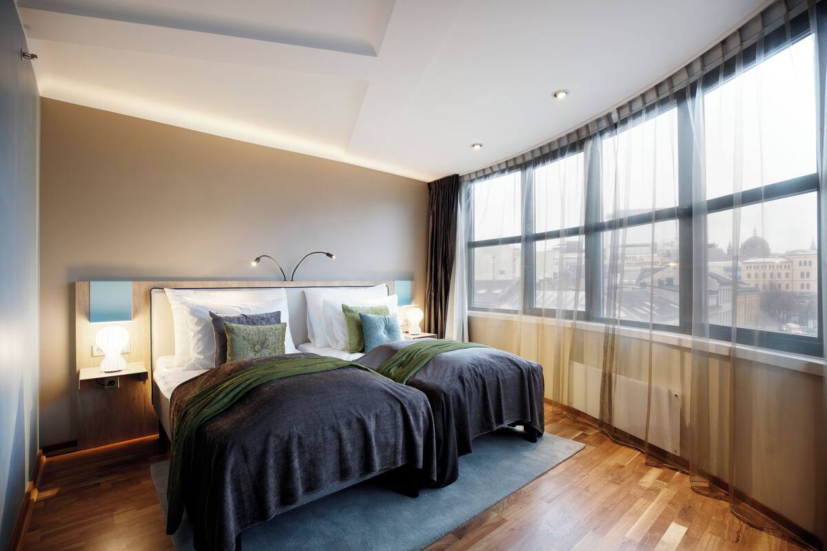 A modern bedroom with a large window, a bed with dark bedding, and a nightstand with a lamp.
