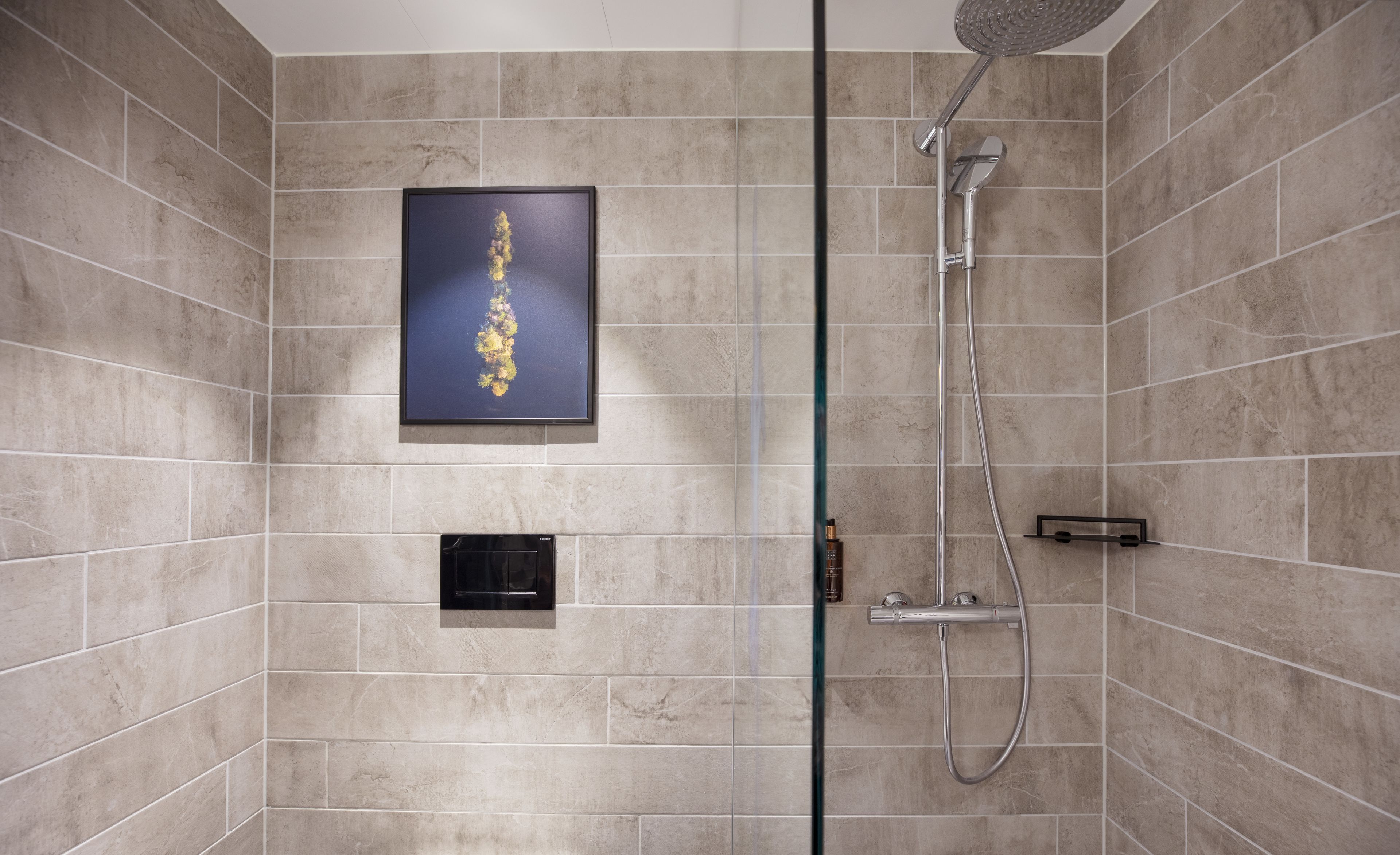 A modern bathroom with beige tiles, a framed artwork, a glass shower enclosure, and a control panel.