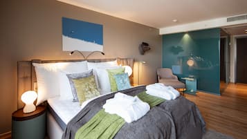 Una habitación de hotel moderna con una cama grande, una mesita de noche con lámpara, una obra de arte en la pared y una zona de estar con una silla y una mesita auxiliar.