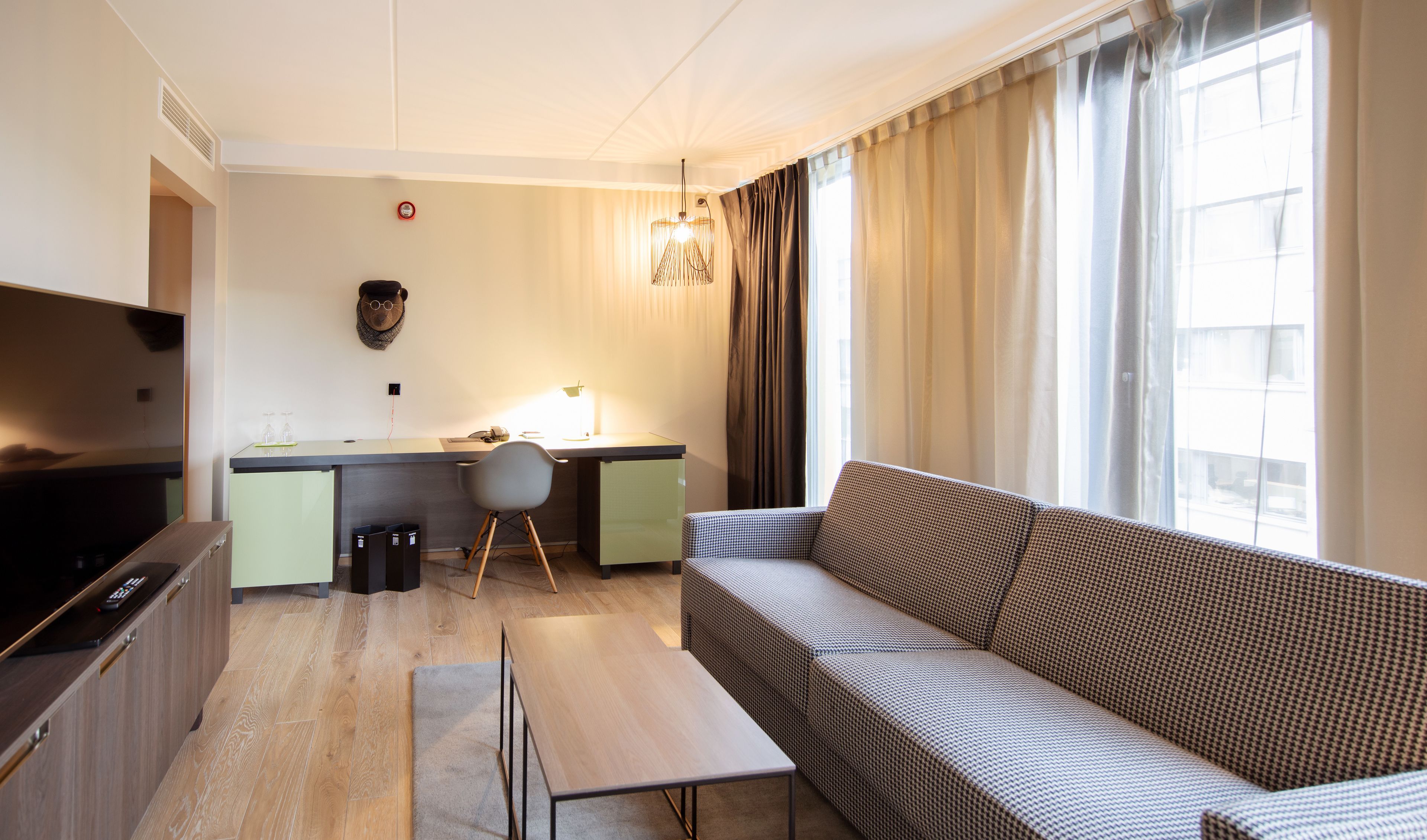 Une chambre d’hôtel moderne équipée d’un canapé, d’un bureau, d’une télévision et d’une grande fenêtre avec des rideaux.