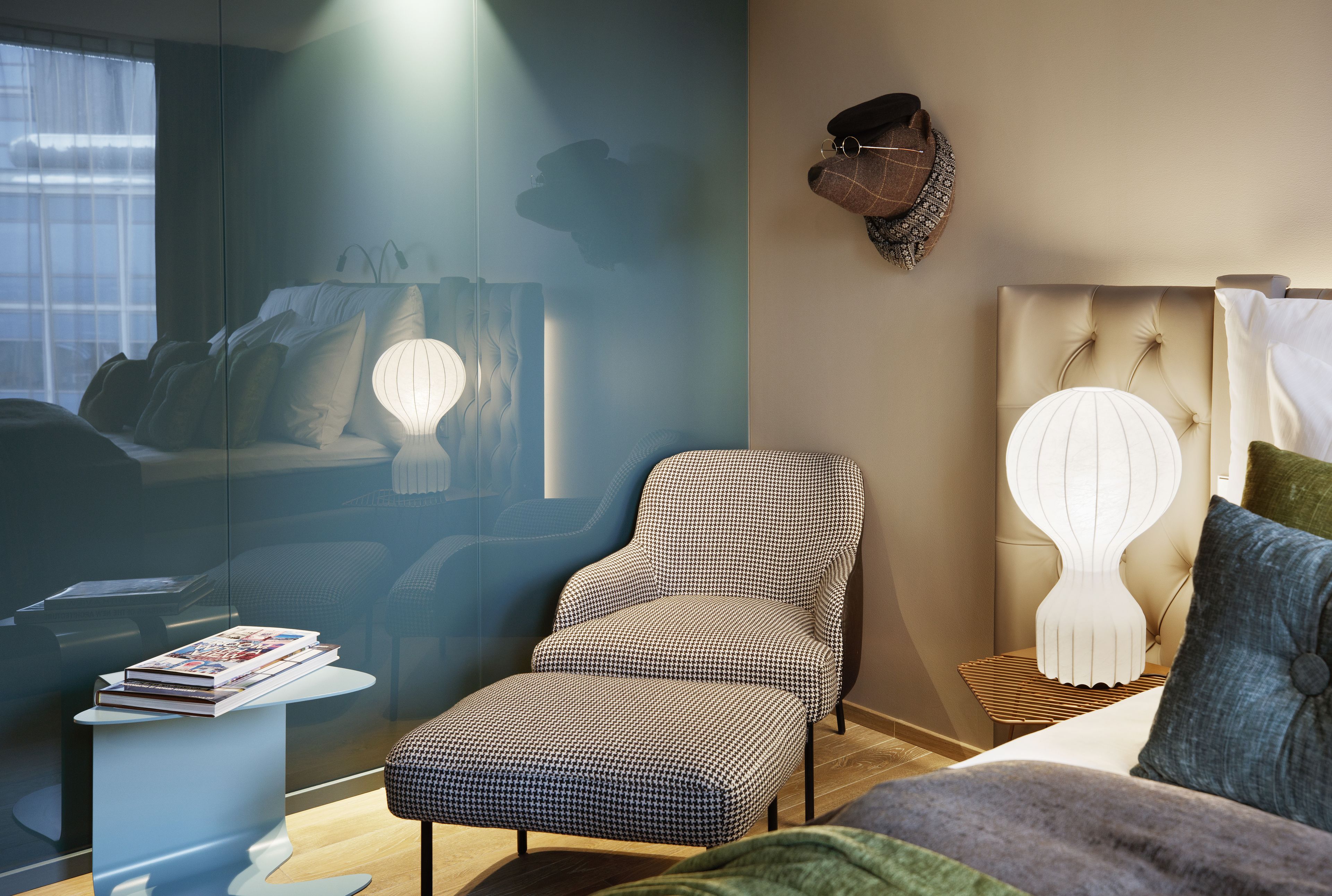 Una habitación de hotel moderna con sofá, una silla, una mesita de noche con lámpara y cabecero fijado a la pared.