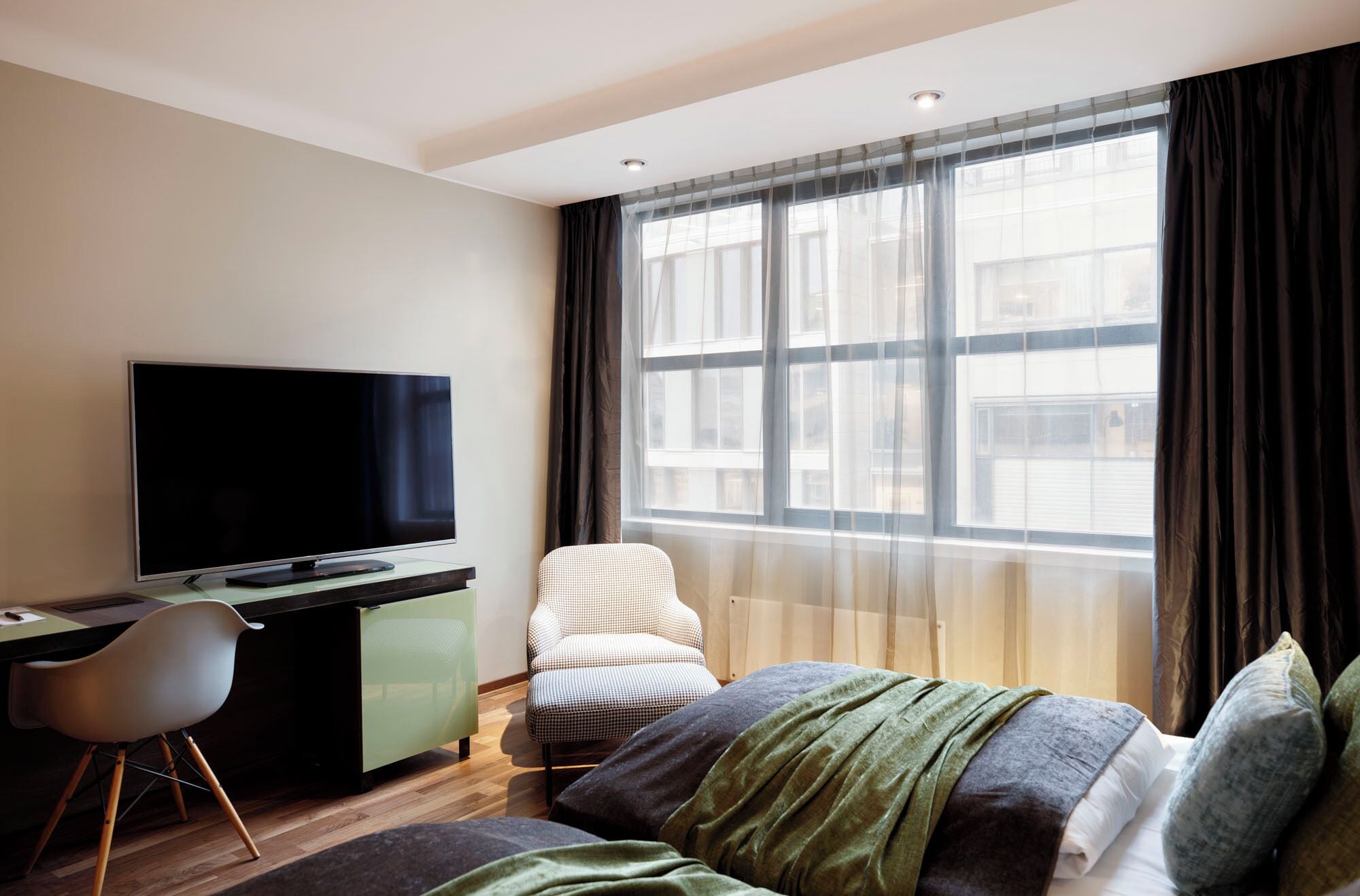 Ein modernes Hotelzimmer mit Flachbildfernseher, Schreibtisch, Stuhl und einem großen Fenster mit Vorhängen.