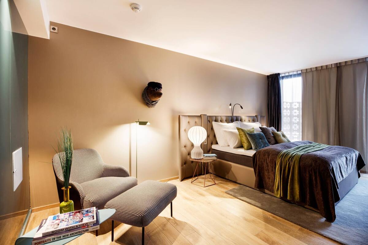 A modern hotel room with a bed, a chair, a small table, and a wall-mounted lamp.