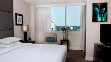 A hotel room with a large bed, a chair, a desk with a lamp, a TV, and a window with a city view.