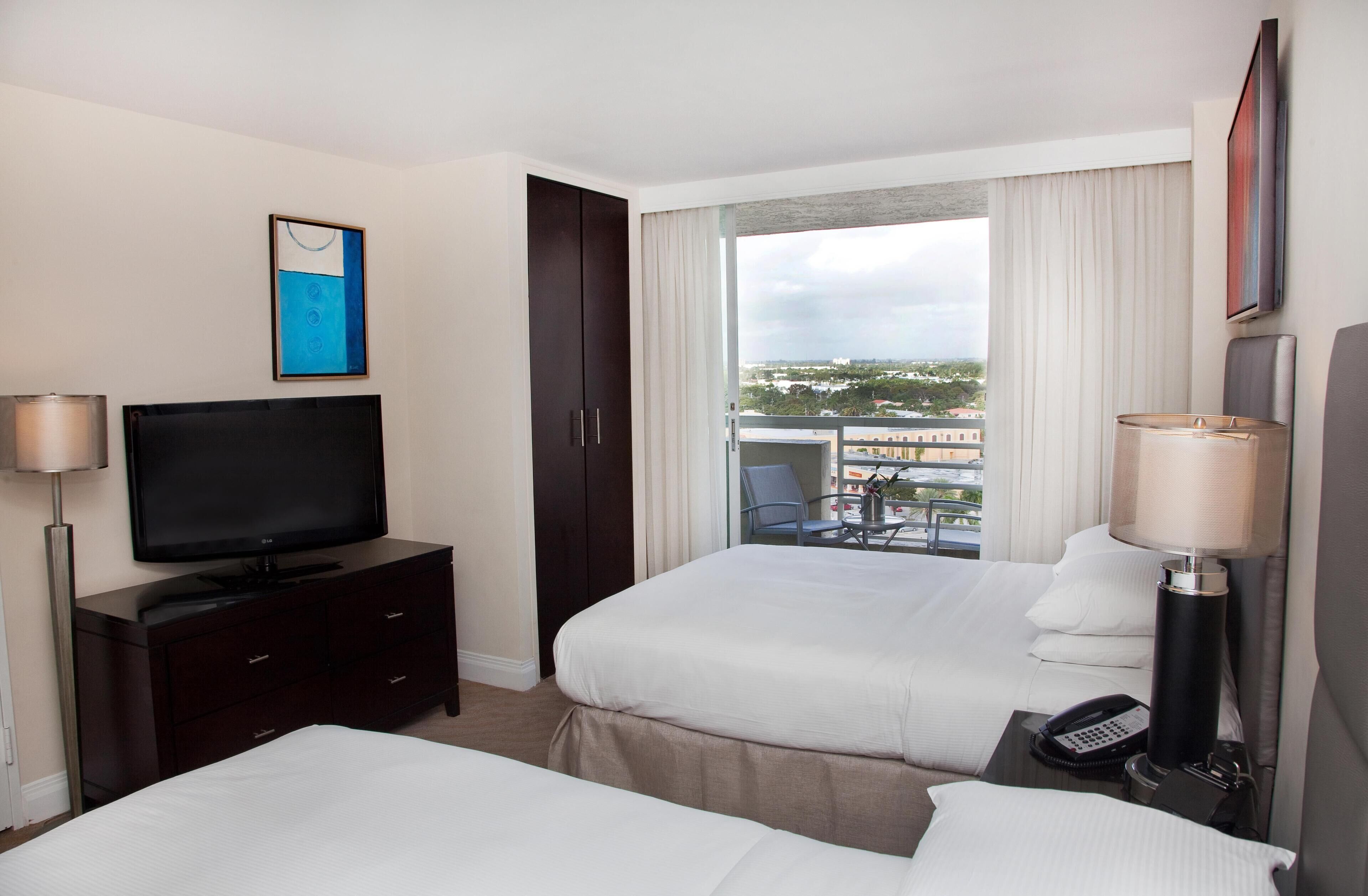 A hotel room with two beds, a TV, a desk with a lamp, a phone, and a balcony with a view of the city.