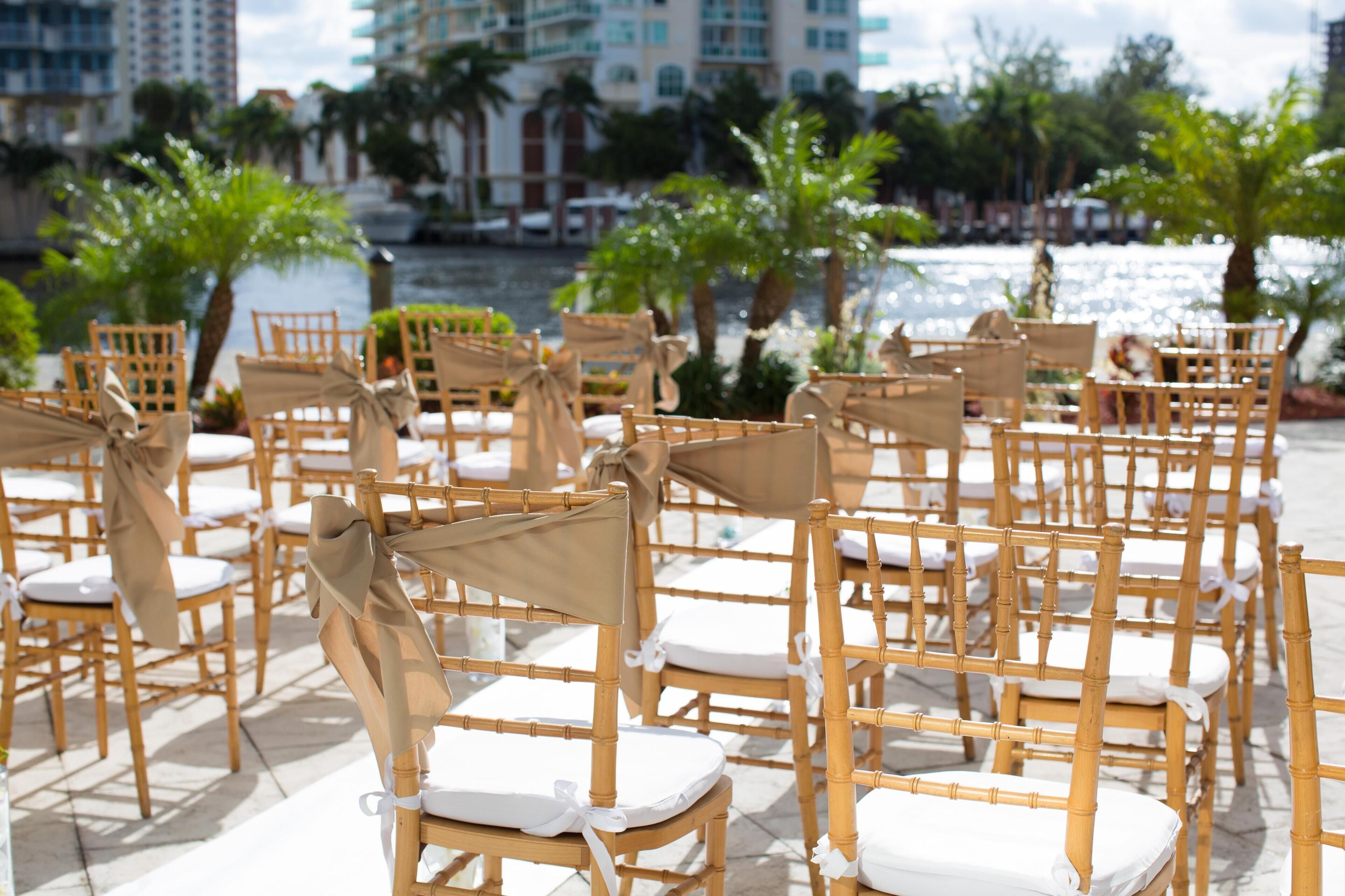 Rows of wooden chairs with white cushions and beige covers, arranged outdoors with a view of a body of water and palm trees in the background.