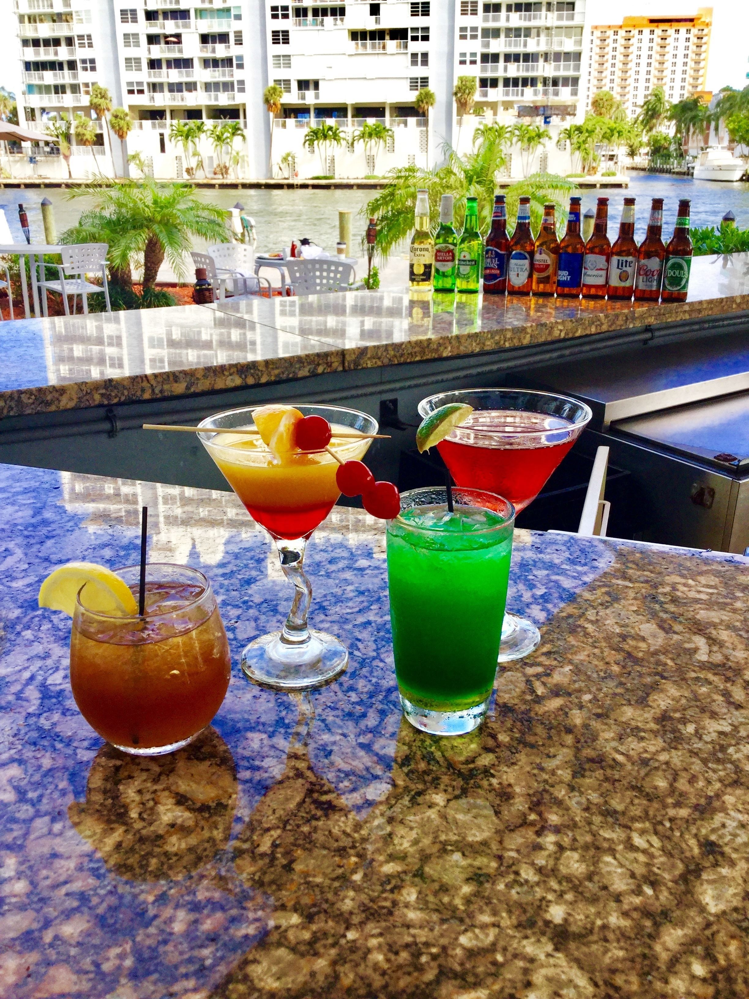 Zona de bar con vistas a un puerto deportivo y edificios. Hay tres bebidas sobre la barra.