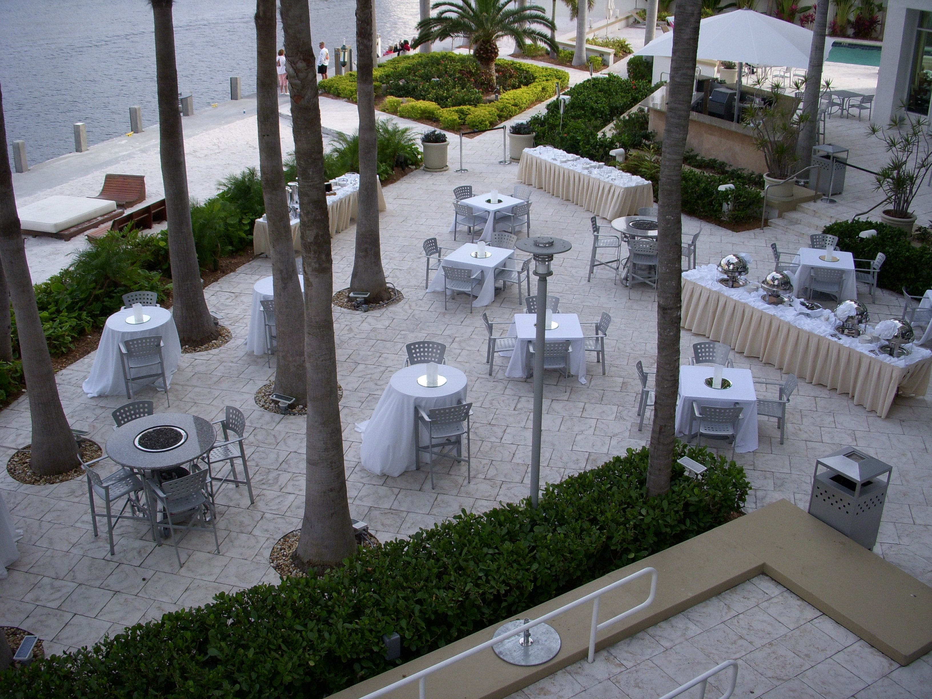 An outdoor dining area with round tables and chairs, surrounded by palm trees and a view of the water.