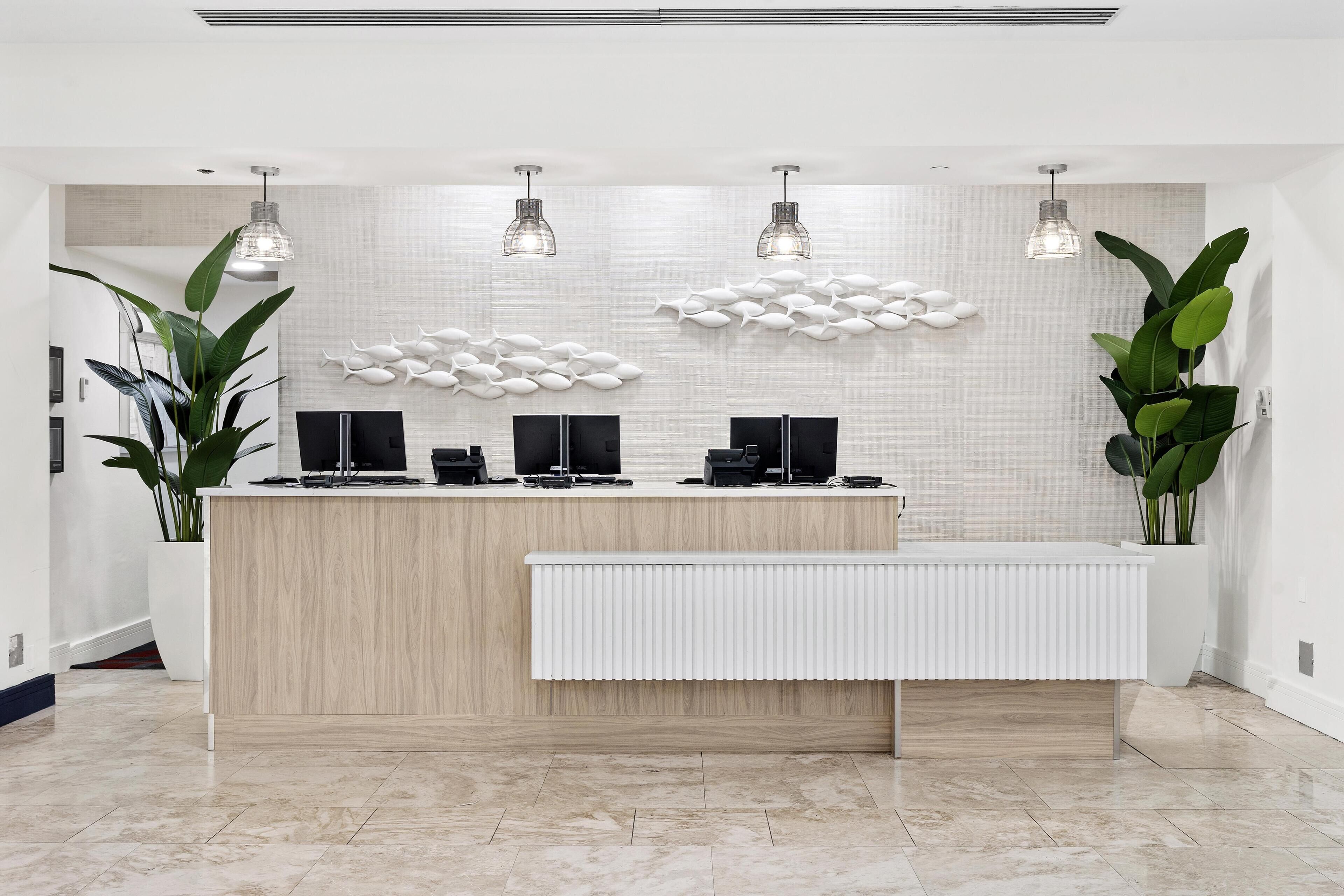 A reception desk with a light wood finish, two black monitors, and a white radiator. There are three pendant lights above the desk, and a wall-mounted fish decoration.