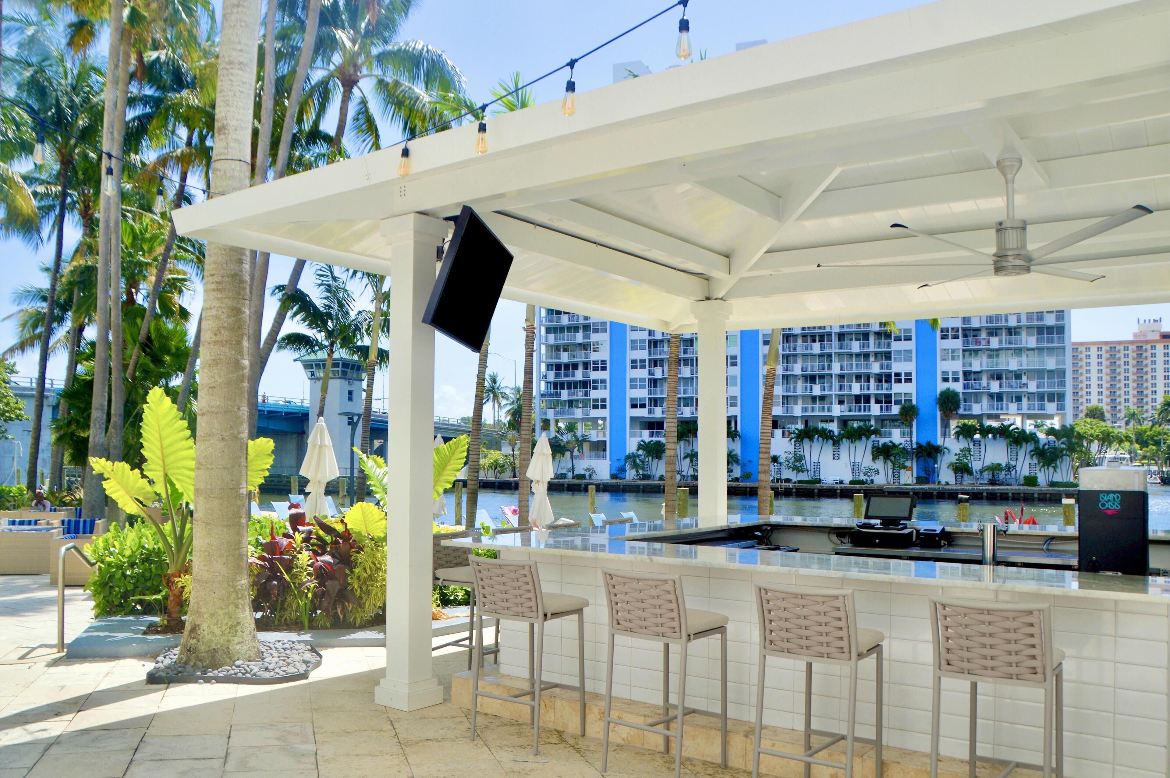 A covered outdoor bar area with a pool view, bar stools, and a TV screen.