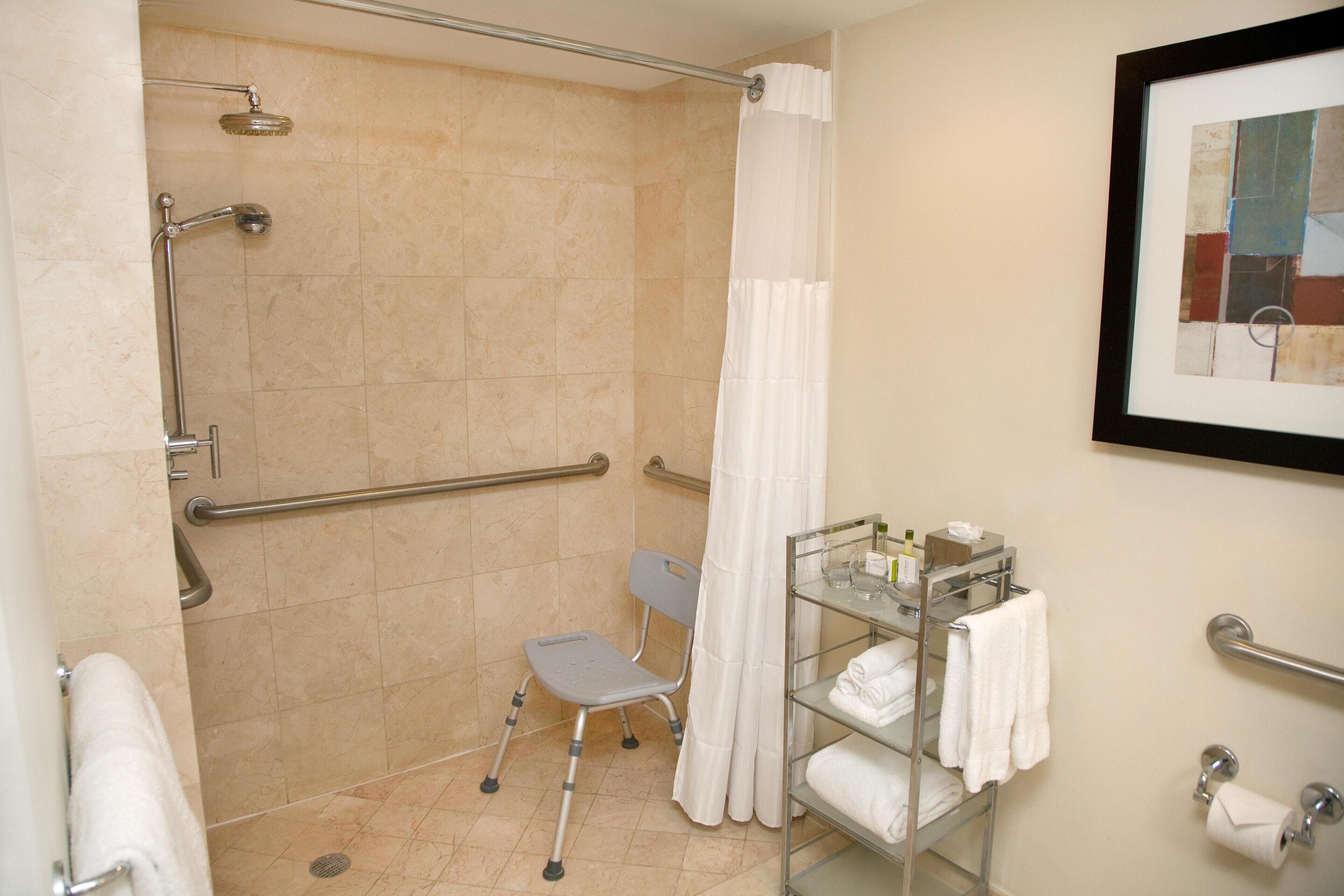 A bathroom with a shower, grab bars, a wheelchair-accessible bench, a towel rack, and a framed picture on the wall.