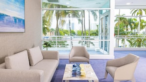 Une chambre d’hôtel moderne dotée d’une grande fenêtre offrant une vue sur des palmiers et une marina. On y trouve un canapé beige, deux fauteuils et une table basse agrémentée d’un objet décoratif.