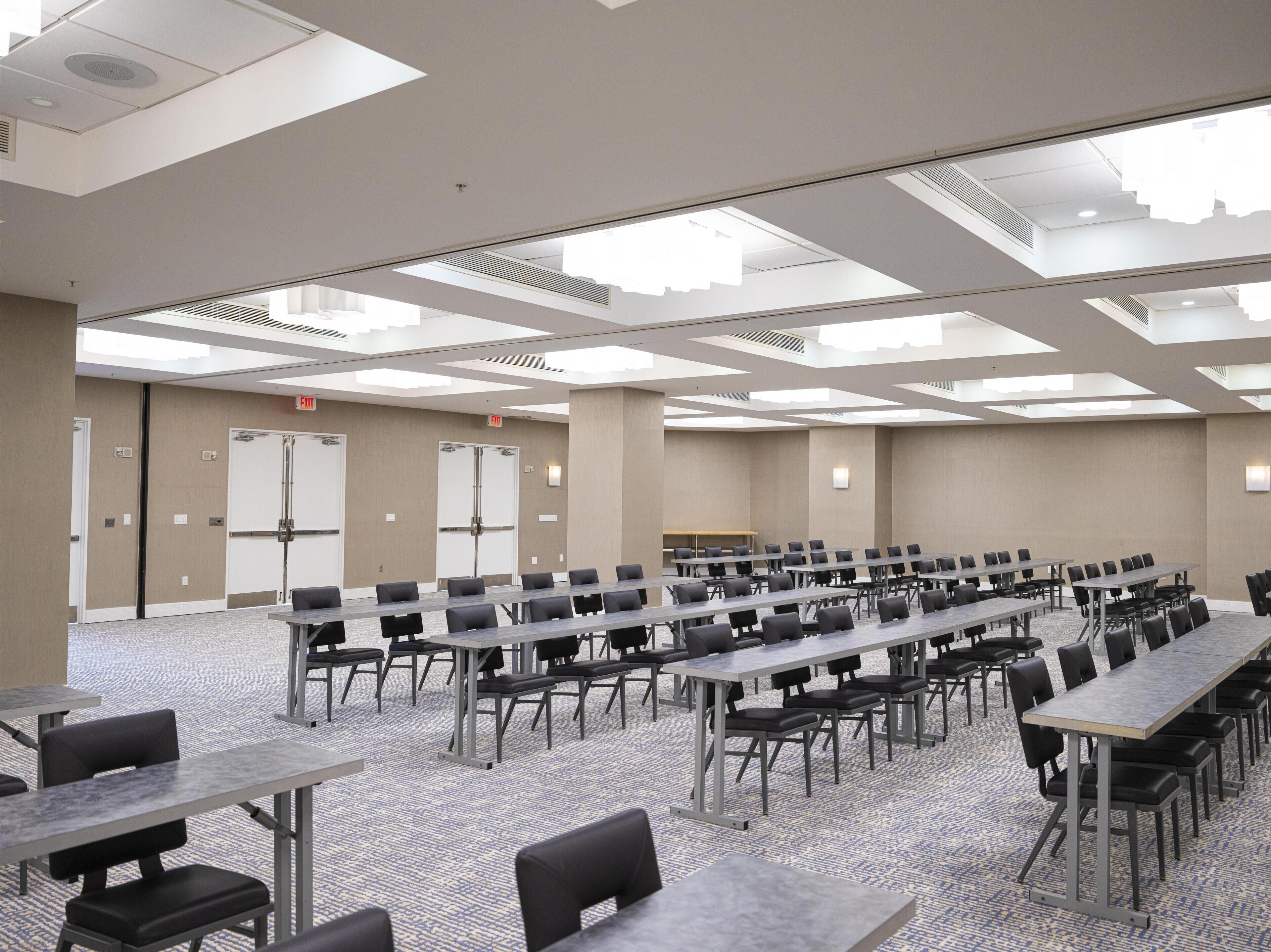 A conference room with multiple tables and chairs, large windows, and a carpeted floor.
