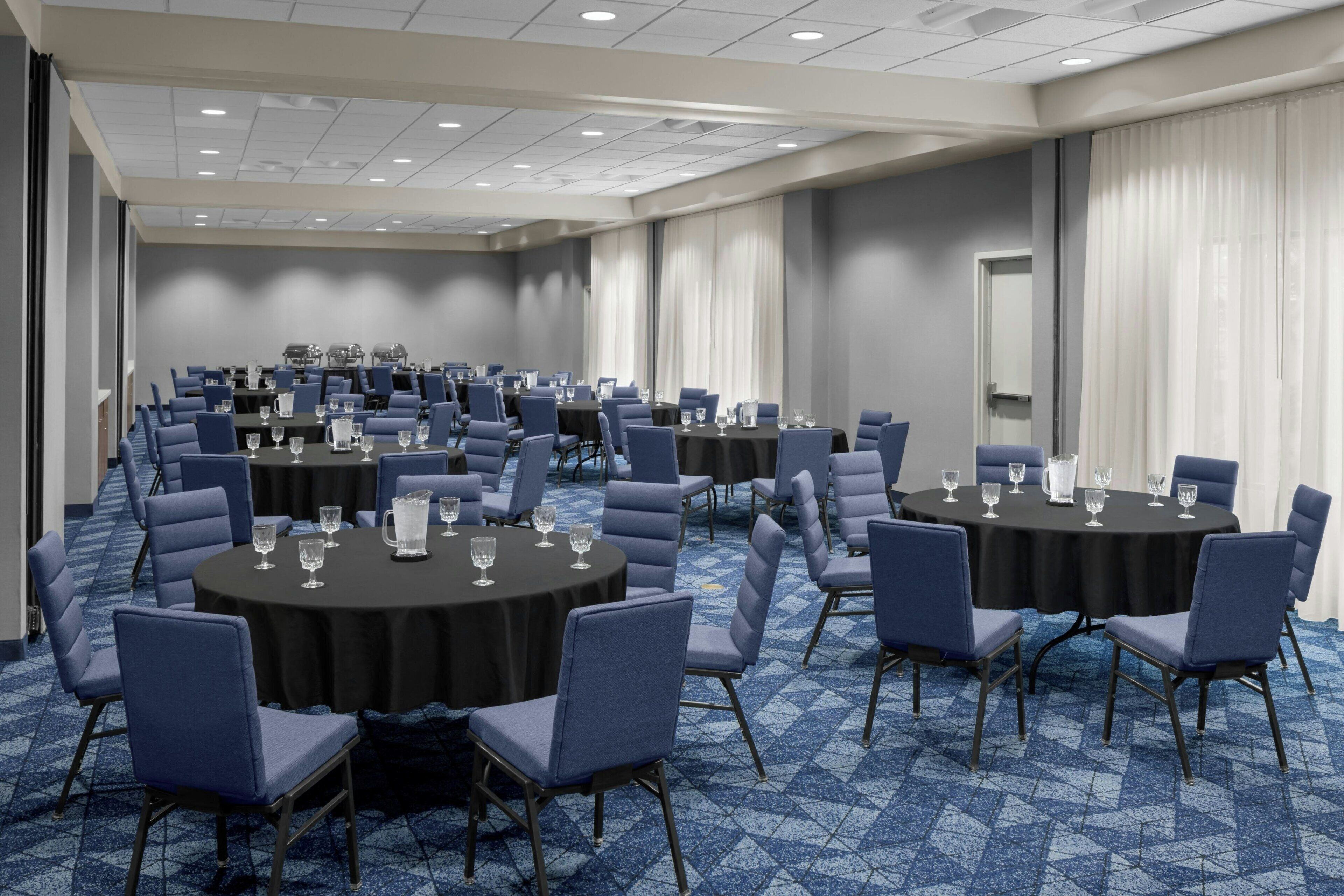 Een ruime congreszaal met meerdere ronde tafels, elk omringd door blauwe stoelen. De zaal heeft een vloerbedekking en een plafond met inbouwverlichting.