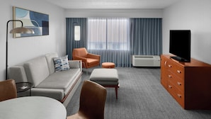 A modern hotel room with a grey sofa, an orange armchair, a small round table, a television on a wooden cabinet, and a large window with curtains.