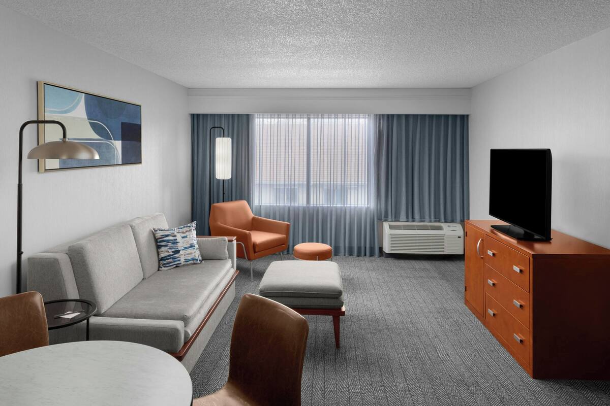 A modern hotel room with a grey sofa, an orange armchair, a small round table, a television on a wooden cabinet, and a large window with curtains.
