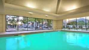 An indoor swimming pool with clear glass windows, a tiled floor, and a view of outdoor seating and a mountain in the background.