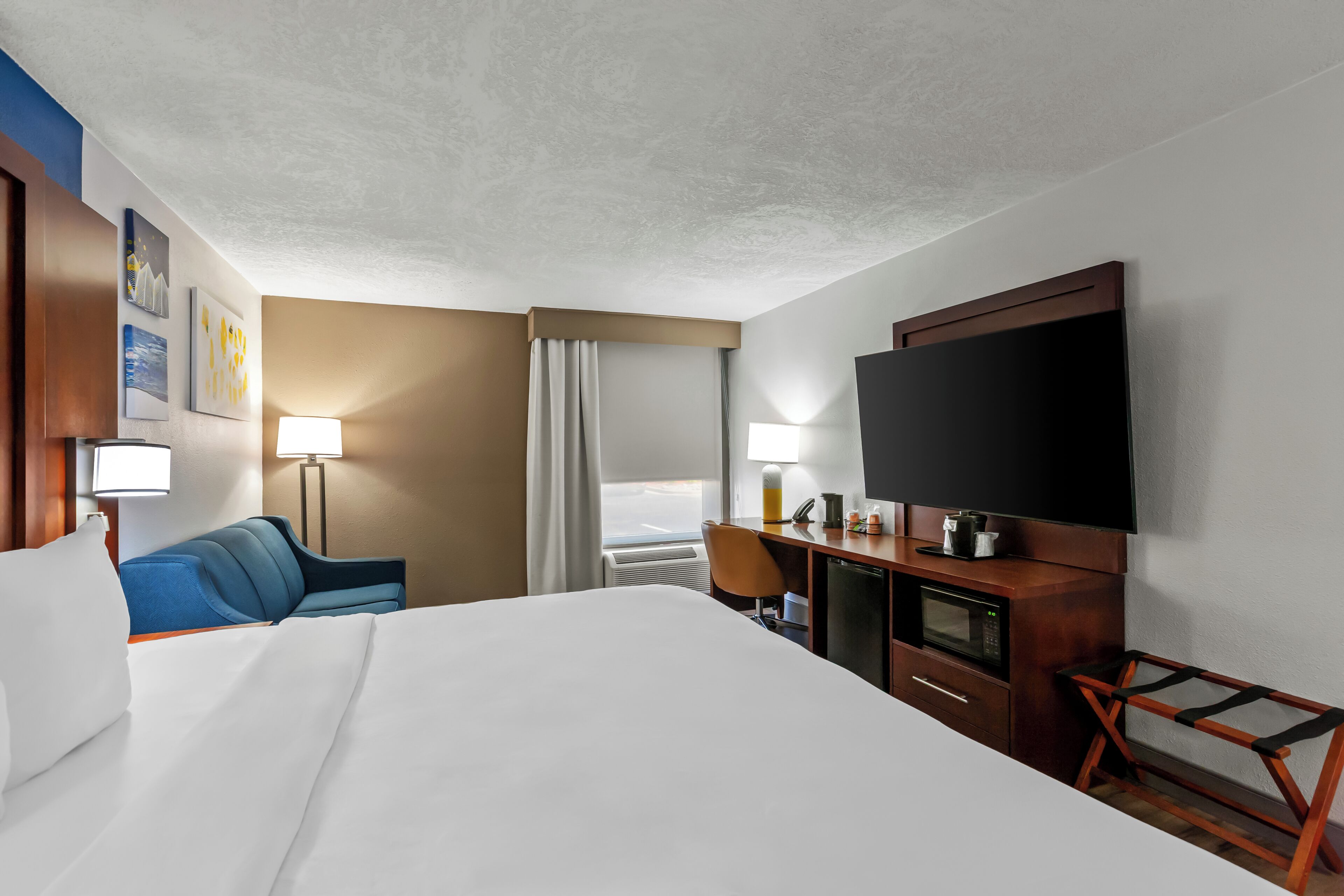 Una habitación de hotel con cama, escritorio, televisor, silla, lámpara y ventana con cortinas.
