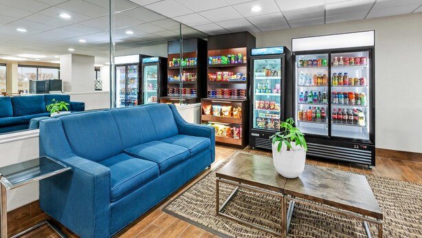 A modern hotel lobby with a blue sofa, a coffee table, a refrigerator stocked with beverages, and a seating area with a view.