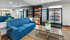 A modern hotel lobby with a blue sofa, a coffee table, a refrigerator stocked with beverages, and a seating area with a view.