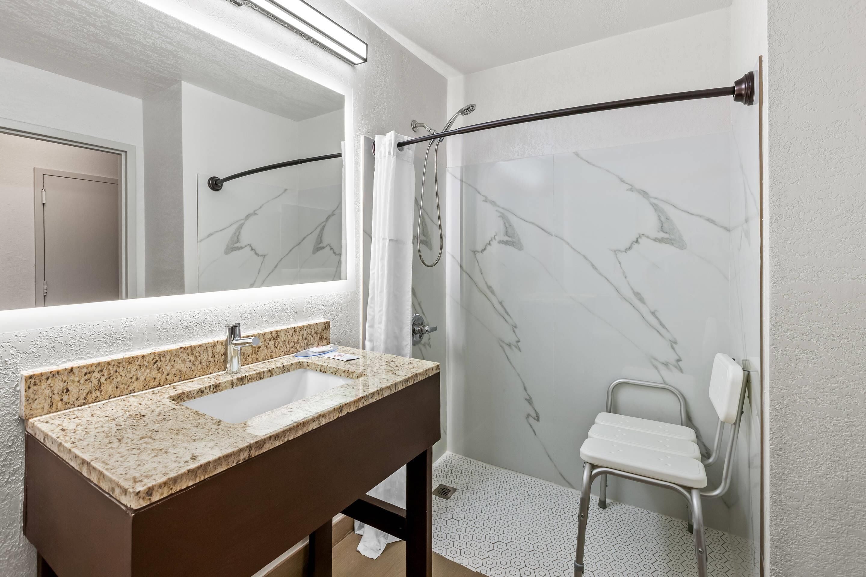 A bathroom with a marble shower wall, a white shower chair, a sink with a granite countertop, and a mirror above the sink.