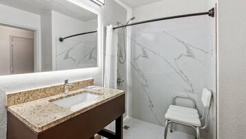A bathroom with a marble shower wall, a white shower chair, a sink with a granite countertop, and a mirror above the sink.