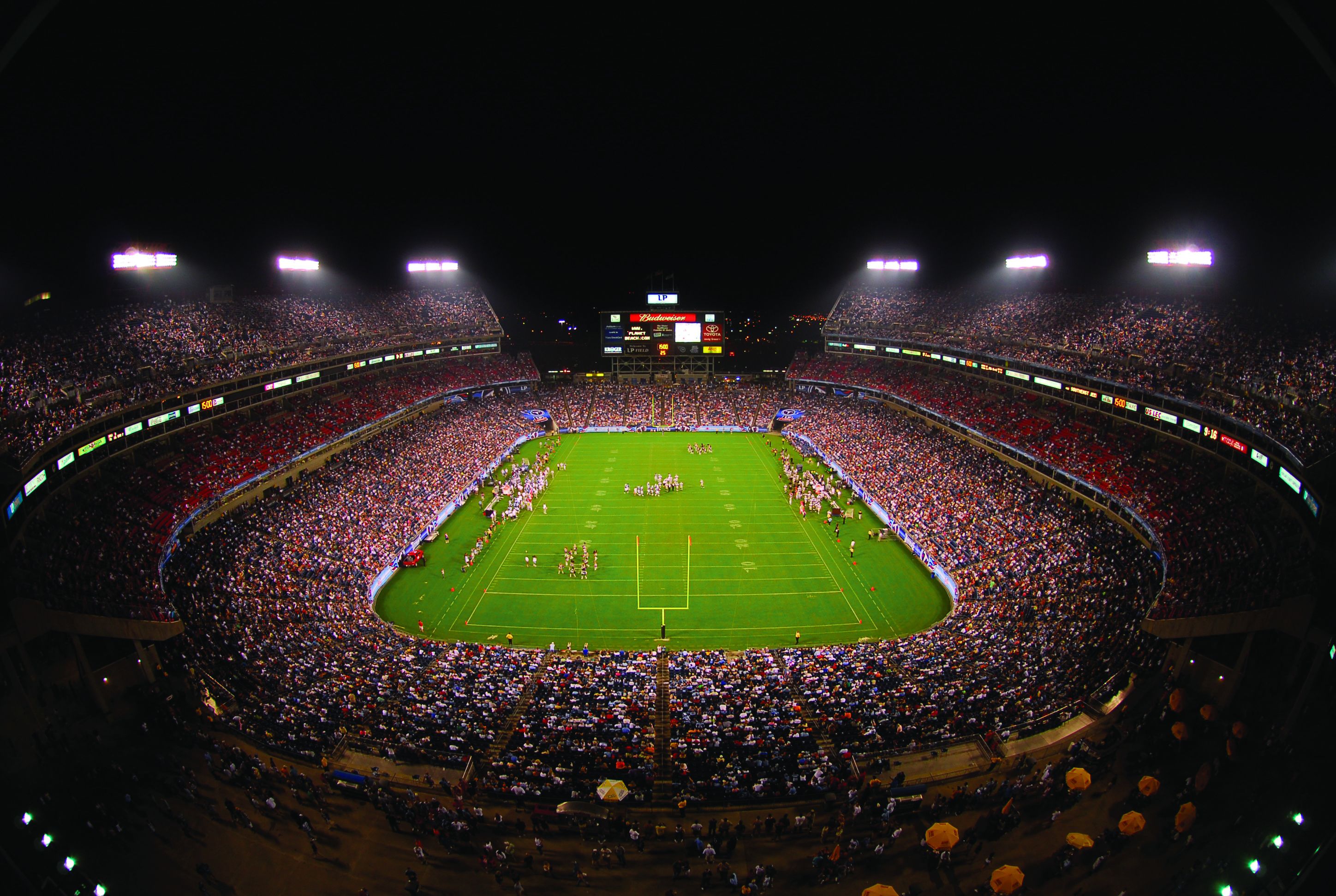 A large stadium with multiple tiers of seating, a central field, and a scoreboard.