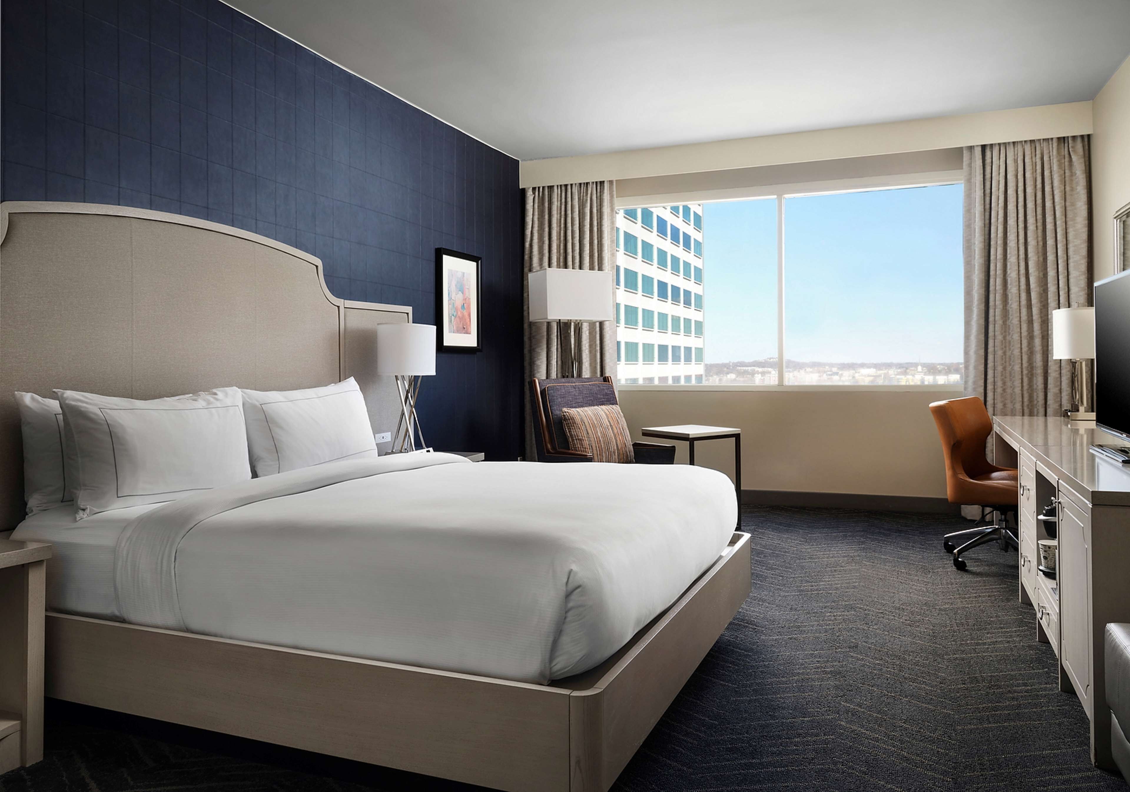 A hotel room with a large bed, a desk, a chair, a lamp, and a view of a cityscape through a large window.