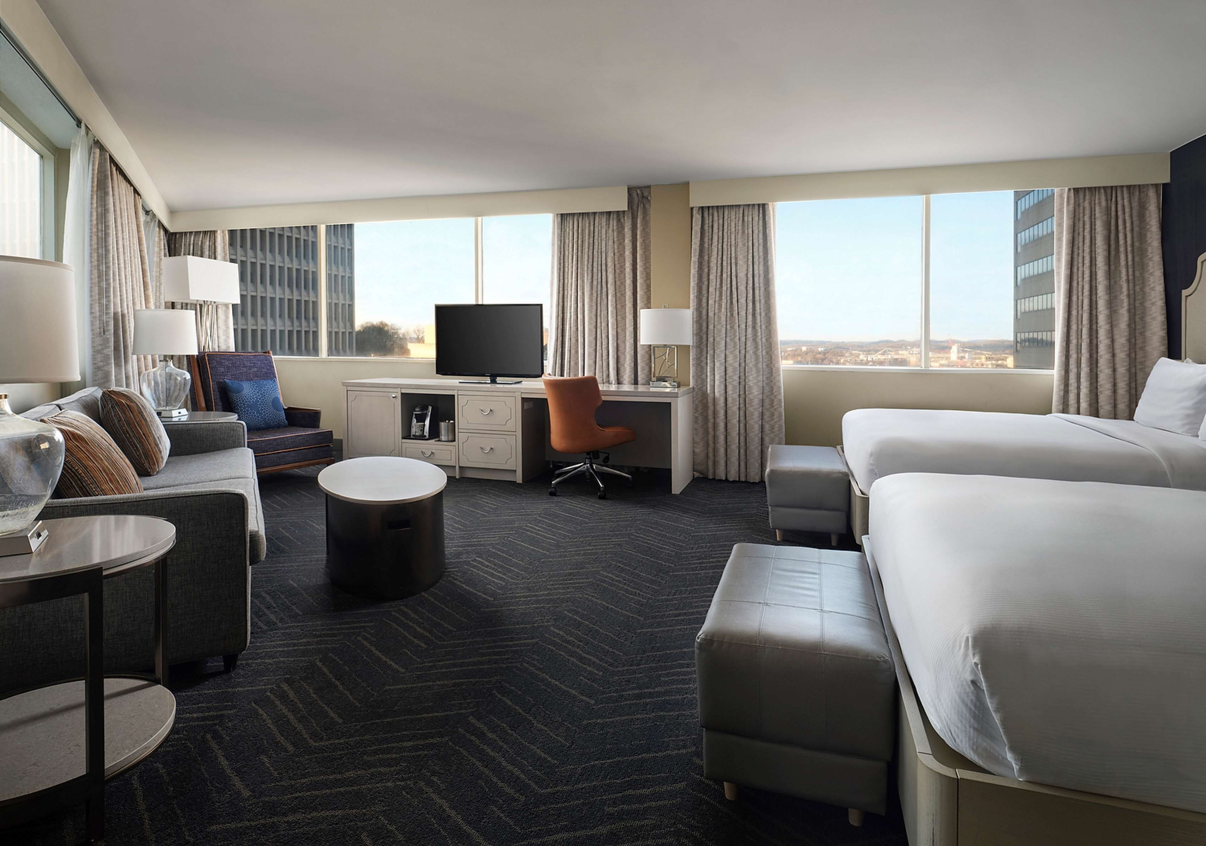 A hotel room with a large bed, a desk with a computer, a chair, a small table, and a view of the city.
