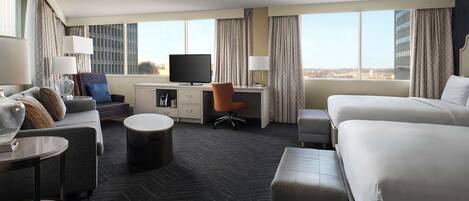 A hotel room with a large bed, a desk with a computer, a chair, a small table, and a view of the city.