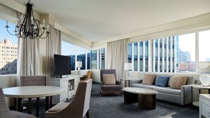Quarto de hotel com uma grande janela, um candelabro, um sofá, uma mesa de centro, uma TV e uma área de jantar.