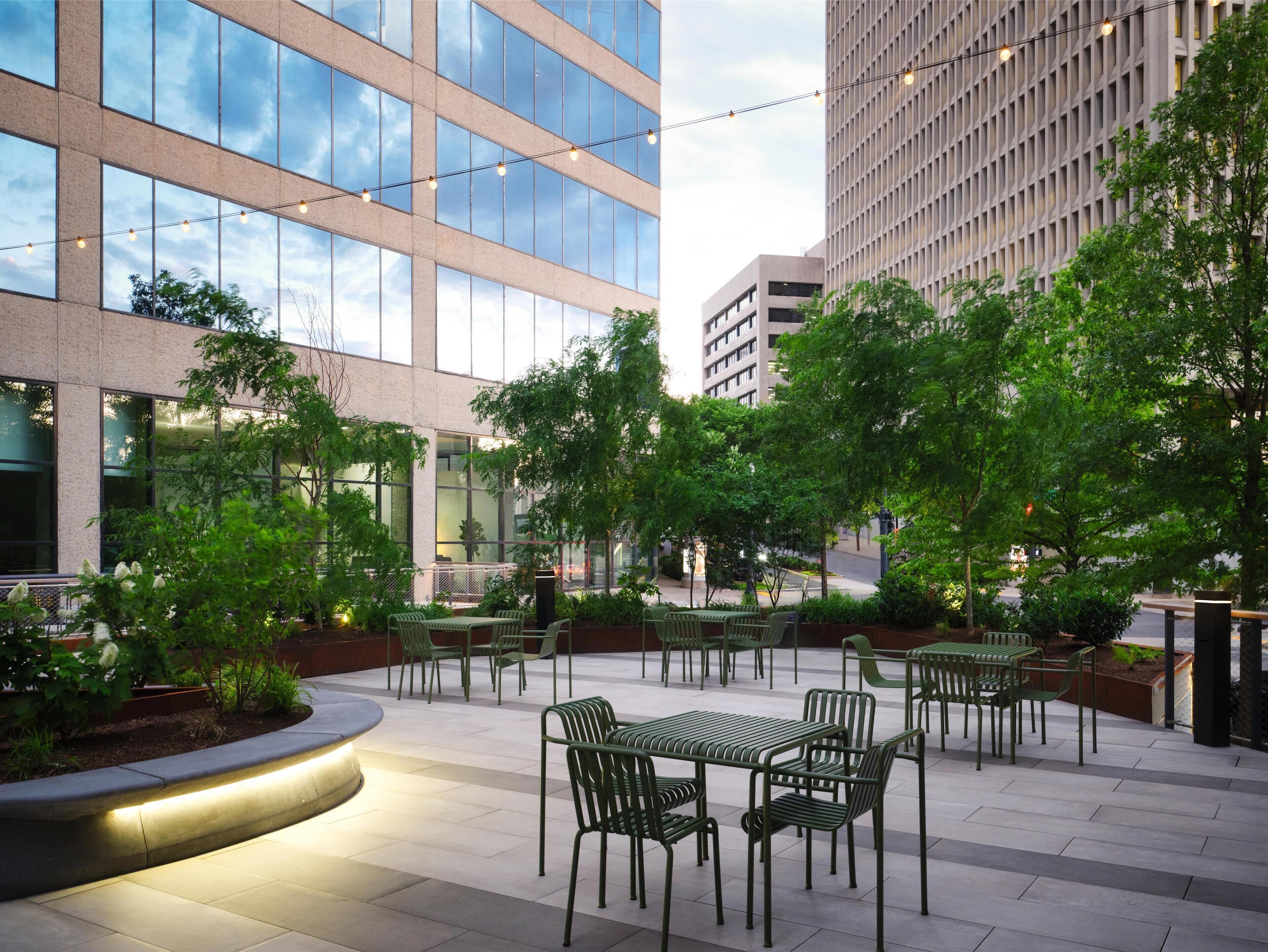An outdoor patio area with green metal tables and chairs, surrounded by trees and a modern building with large windows.
