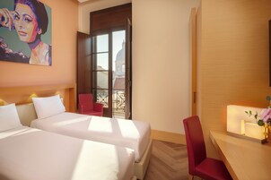 Habitación de hotel con dos camas, una silla roja, un escritorio con lámpara y un cuadro en la pared.