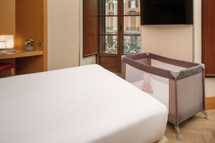 Une chambre d’hôtel avec un lit, un bureau doté d’une lampe, une petite plante et une fenêtre donnant sur des immeubles.