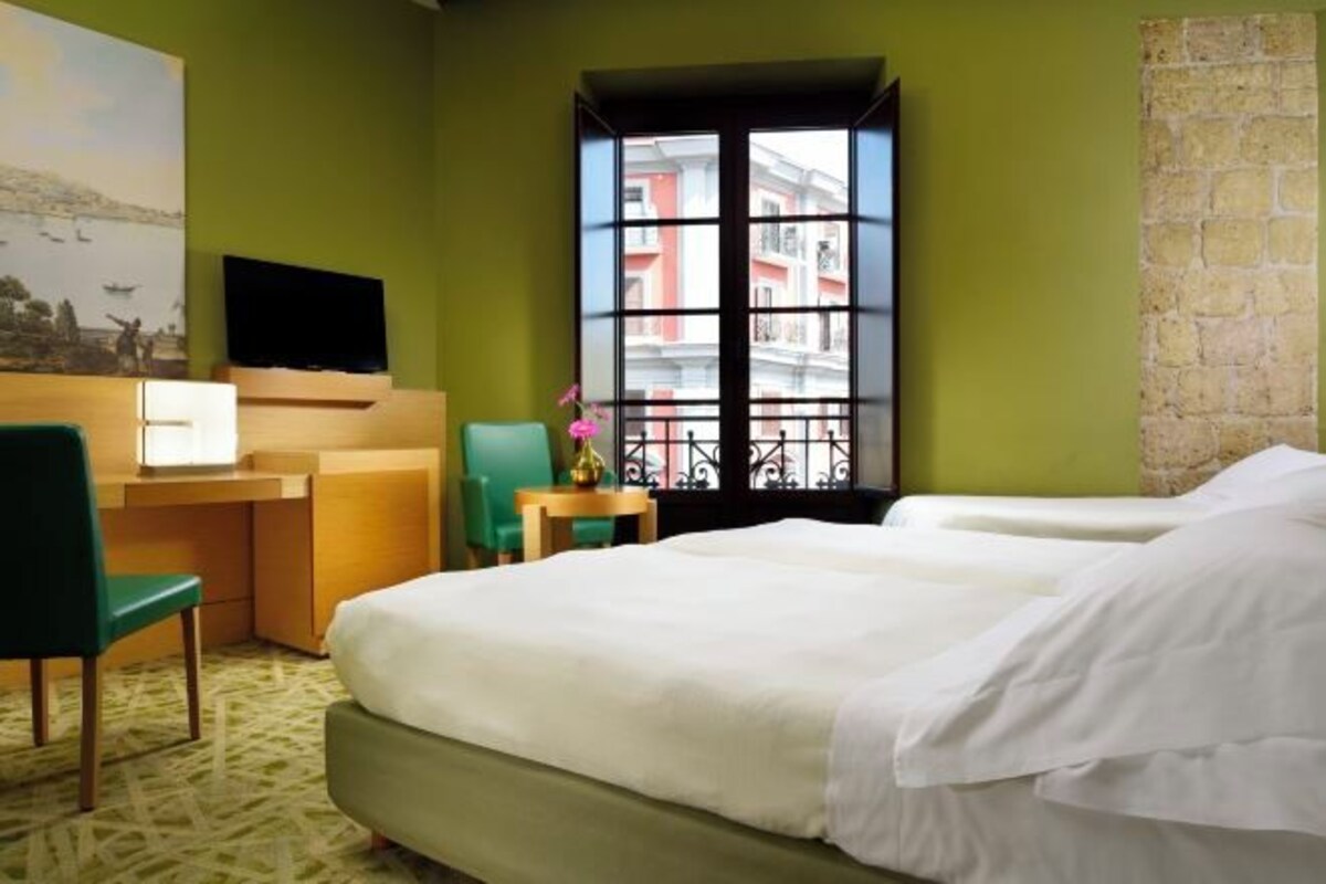 A hotel room with a bed, a desk, a chair, a TV, and a window with a view of buildings.