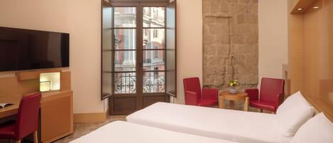 A hotel room with two beds, a TV, a desk with a chair, a small table with a plant, and a large window with a view of buildings.