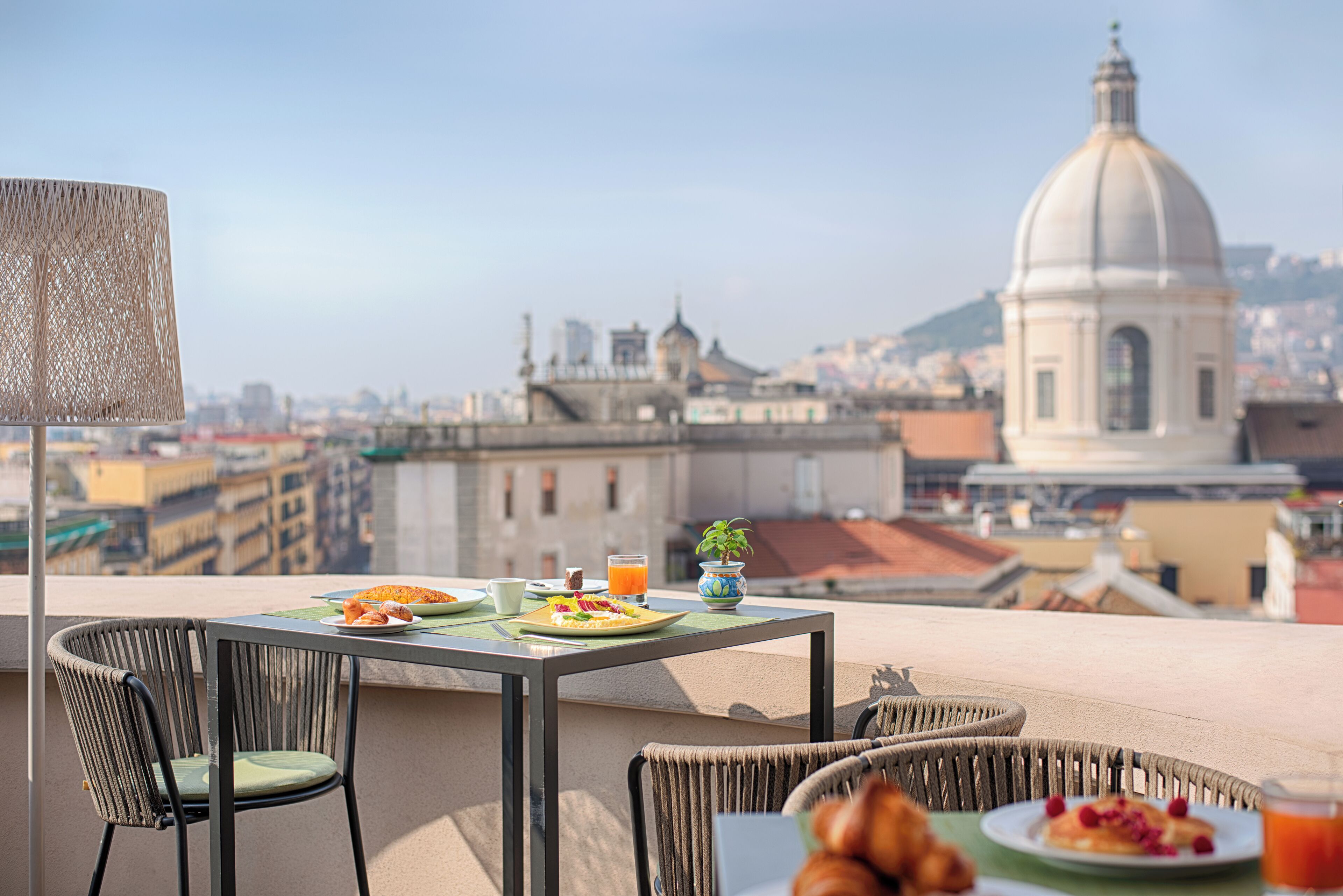Una mesa de comedor con platos de comida y bebidas, sillas de mimbre y vista a un paisaje urbano con un edificio de forma abovedada.