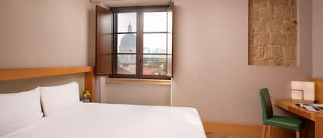 Una habitación de hotel con una cama grande, un escritorio con silla, un ventanal con vista a un edificio y una obra de arte colgada en la pared.