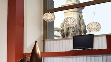 Uma sala de estar moderna com sofá de couro preto, detalhes em vermelho, uma televisão em uma estante branca e uma grande janela com vista para uma estátua e uma fonte.