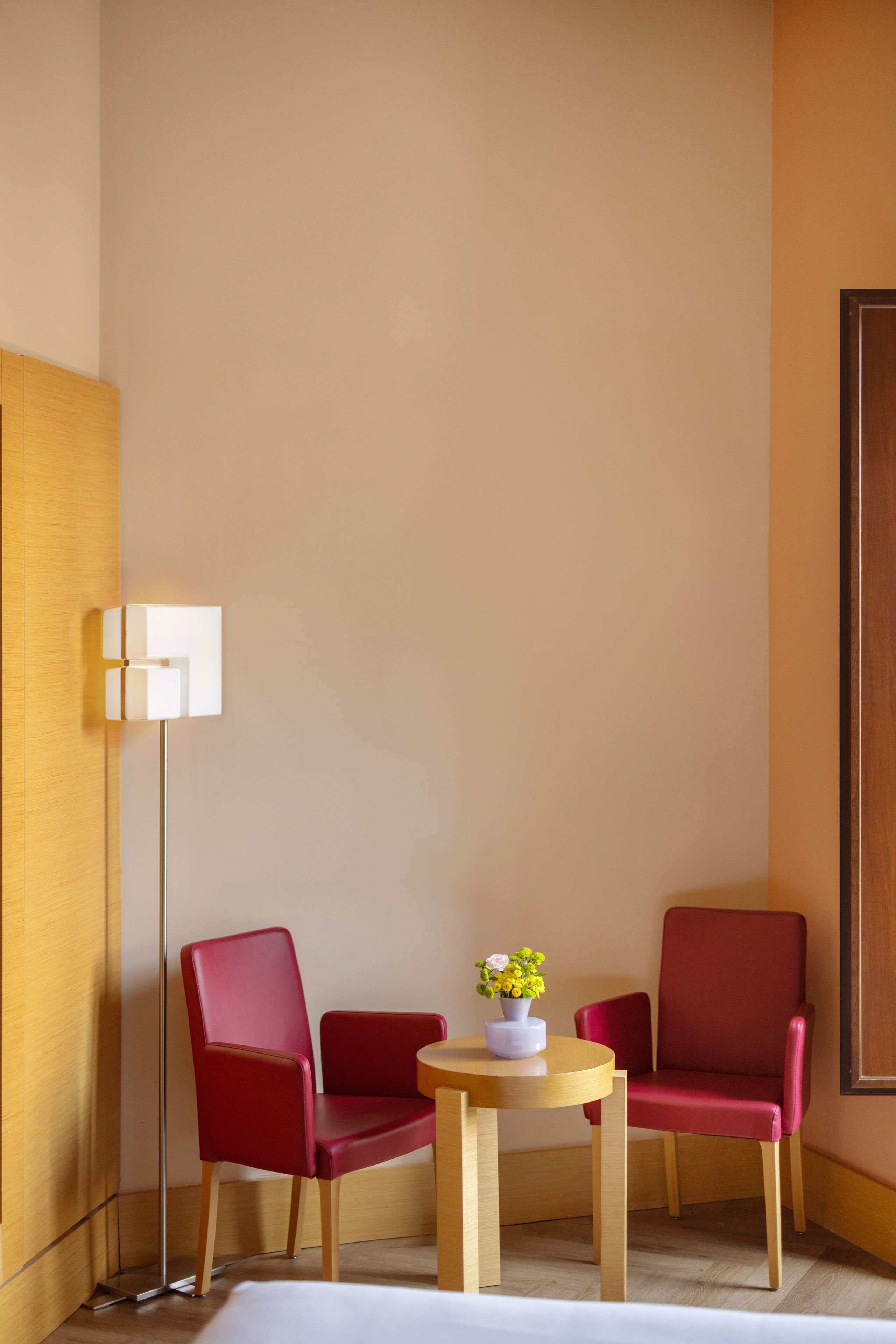 A corner room with two red chairs, a small wooden table with a vase of flowers, and a floor lamp.