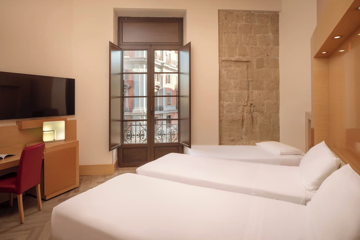 Een hotelkamer met twee bedden, een televisie, een bureau met stoel en een groot raam met uitzicht op gebouwen.