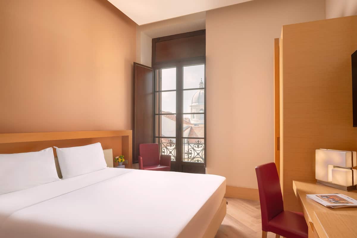 Een hotelkamer met een groot bed, een bureau, een stoel en een raam met uitzicht op gebouwen.