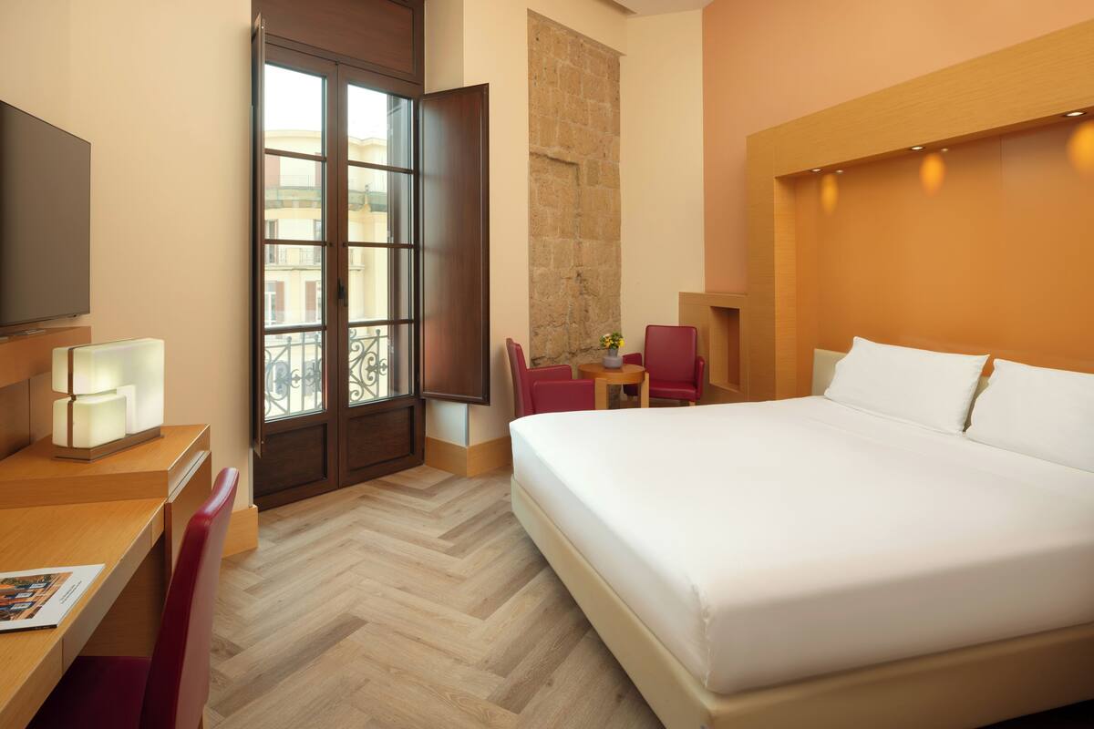 Een hotelkamer met een groot bed, een bureau met stoel, een televisie en een raam met uitzicht op gebouwen.