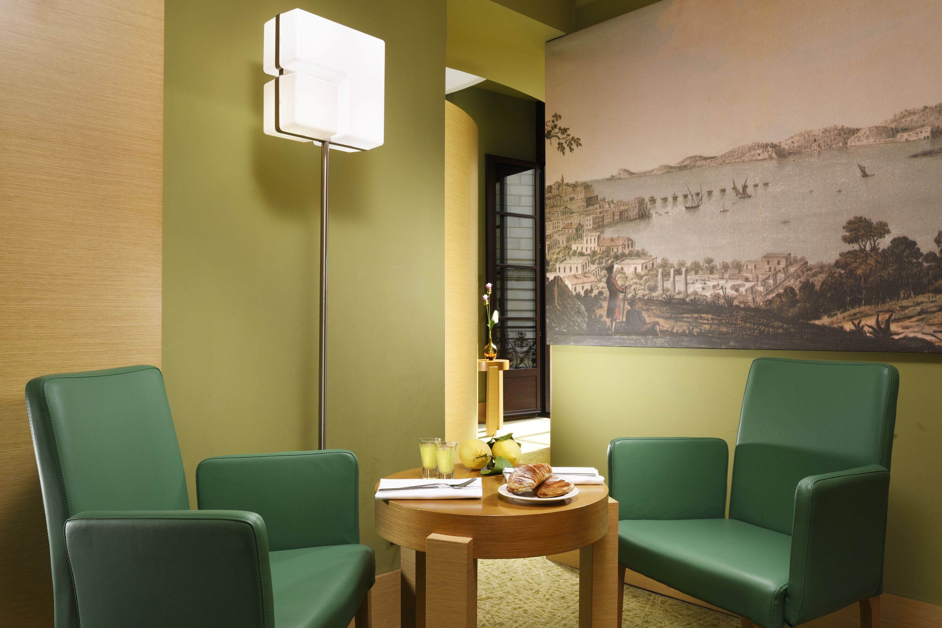 Una habitación con dos sillas verdes, una mesa redonda de madera con alimentos para el desayuno, un cuadro de un pueblo costero y una lámpara de pie.
