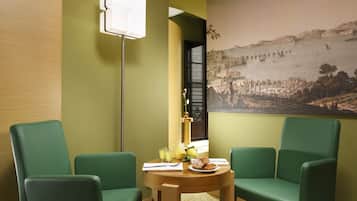 Una habitación con dos sillas verdes, una mesa redonda de madera con alimentos para el desayuno, un cuadro de un pueblo costero y una lámpara de pie.