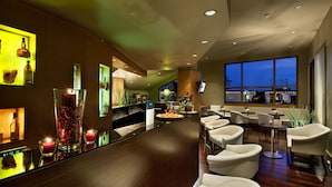 A modern bar area with a long counter, multiple seating options, and a large window providing a view of the outside. - UNA Hotels Napoli (Naples)